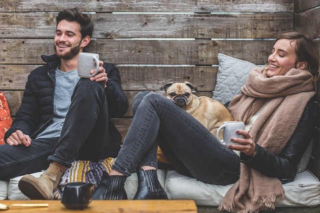 Man and woman lounging on the couch in a cabin. Woman is holding a pug dog and cup of coffee. Very happy.