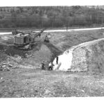 A crane and an old 1950's pick-up truck sitting on top of a pile of dirt above a drain field. Large pipe is sticking sideways out of the drain field with water running out of it into the drain field. Man is standing on the pipe. Two more are observing.