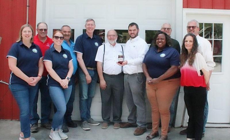 Maryland Department of Agriculture Secretary Kevin Attichs, staff, and representatives of agriculture posing with a jar of jelly in front of a red building.