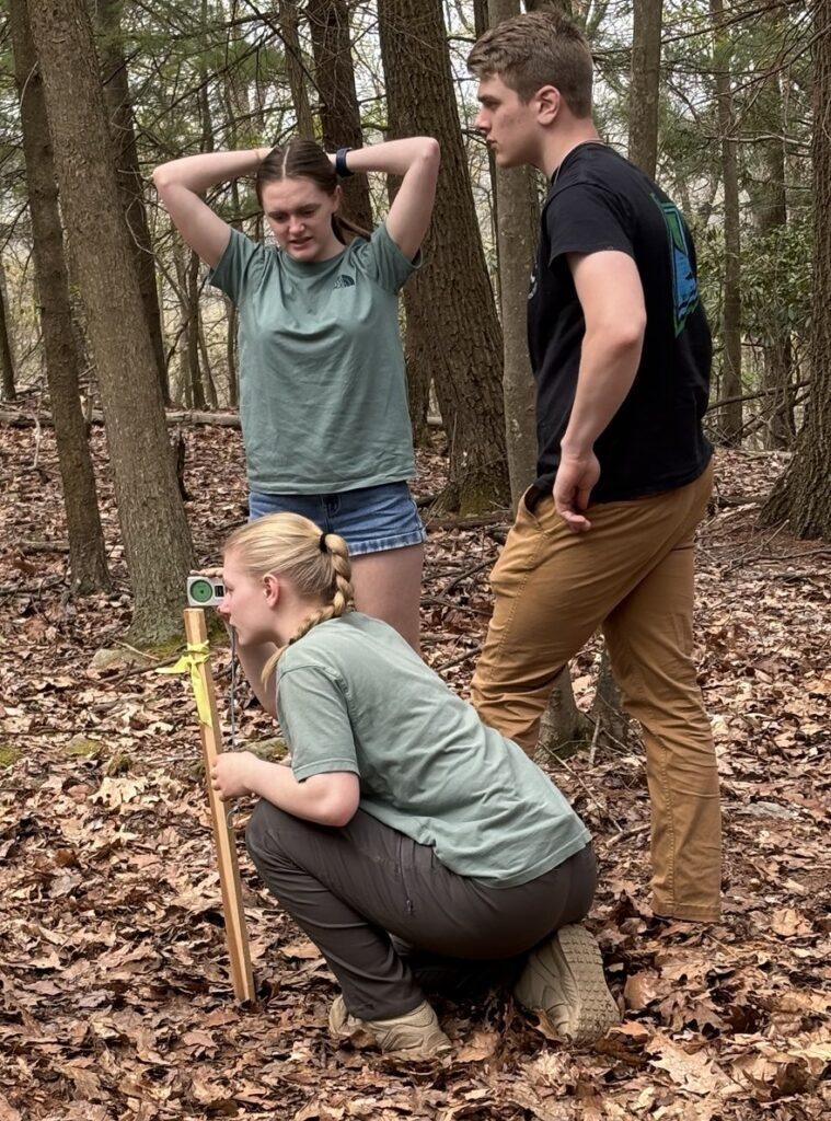 A student squats on the ground looking through survey equipment, while two other students stand over her looking frustrated.