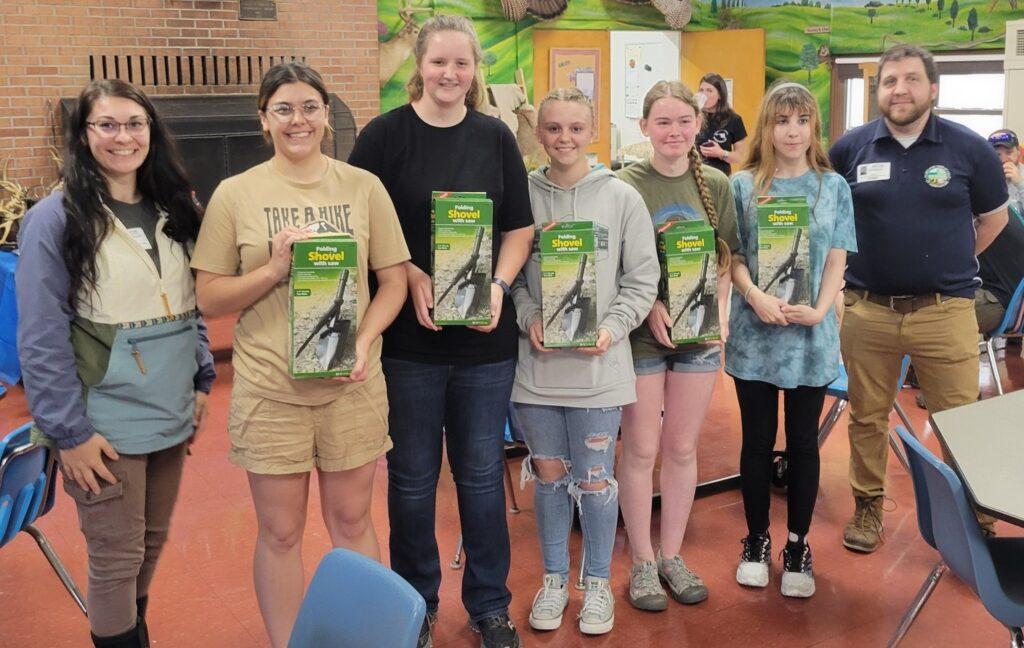 5 Students stand in a row holding boxes containing a shovel. 2 staff members stand on each side.