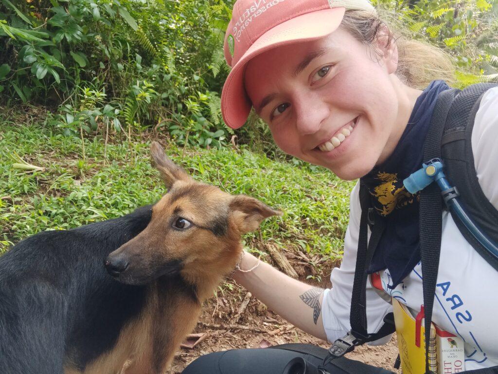 Young woman petting a dog, wearing a red ball cap and carrying a notebook.